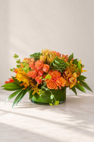 Abundance fall floral arrangement with orange roses, cymbidium orchids, pincushion protea, seasonal greens, and autumn foliage in a glass vase.