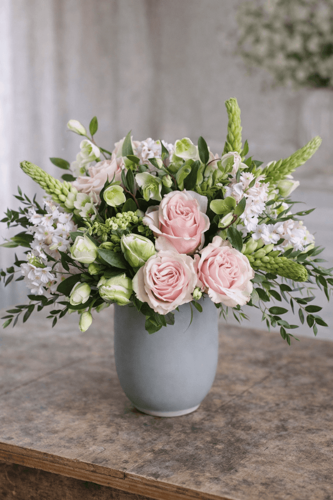 Adore floral arrangement in a matte gray vase with blush roses, white lisianthus, soft lavender blooms, and seasonal greenery.