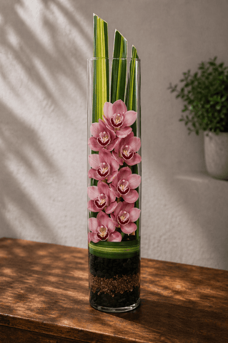 Pink cymbidium orchids arranged vertically in a tall glass cylinder vase with ti leaves and layered stones on a wooden table with soft natural light