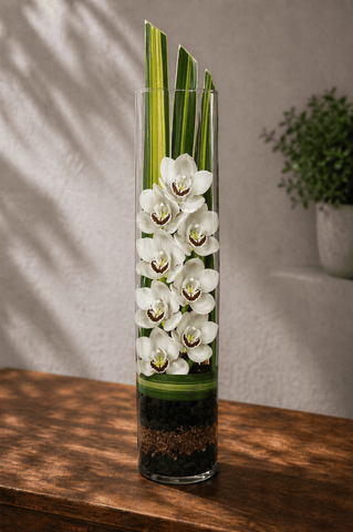 White cymbidium orchids arranged vertically in a tall glass cylinder vase with ti leaves and layered stones on a wooden table with soft natural light
