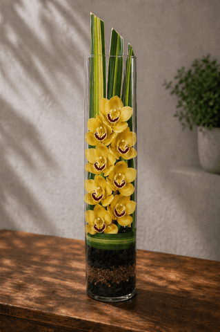 Yellow cymbidium orchids arranged vertically in a tall glass cylinder vase with ti leaves and layered stones on a wooden table with soft natural light