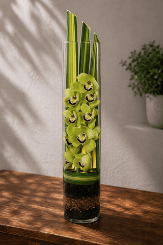 Green cymbidium orchids arranged vertically in a tall glass cylinder vase with ti leaves and layered stones on a wooden table with soft natural light