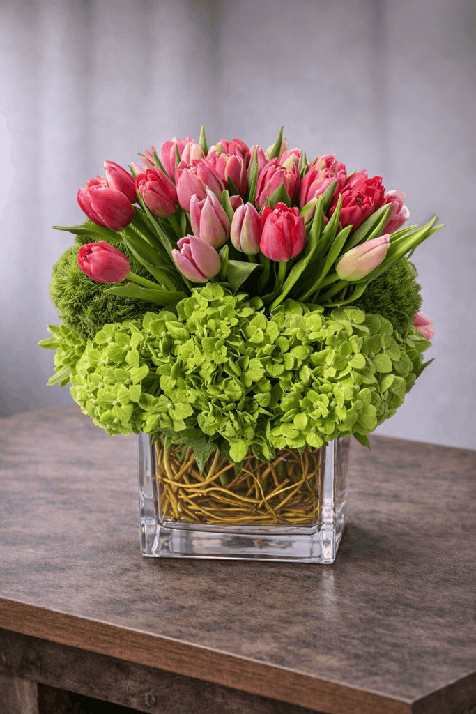 Artful floral arrangement in a clear glass cube vase with pink tulips, green hydrangea, and curly willow branches.