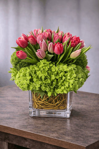 Artful floral arrangement in a clear glass cube vase with pink tulips, green hydrangea, and curly willow branches.
