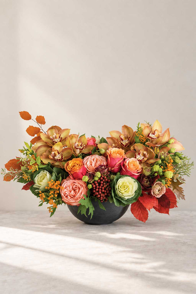Autumn floral arrangement with orange and peach roses, cymbidium orchids, ornamental kale, berries, and fall foliage in a low modern container.