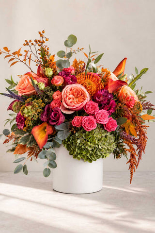 Autumn Muse floral arrangement with coral roses, orange pincushion protea, calla lilies, green hydrangea, and fall foliage in a matte white vase.