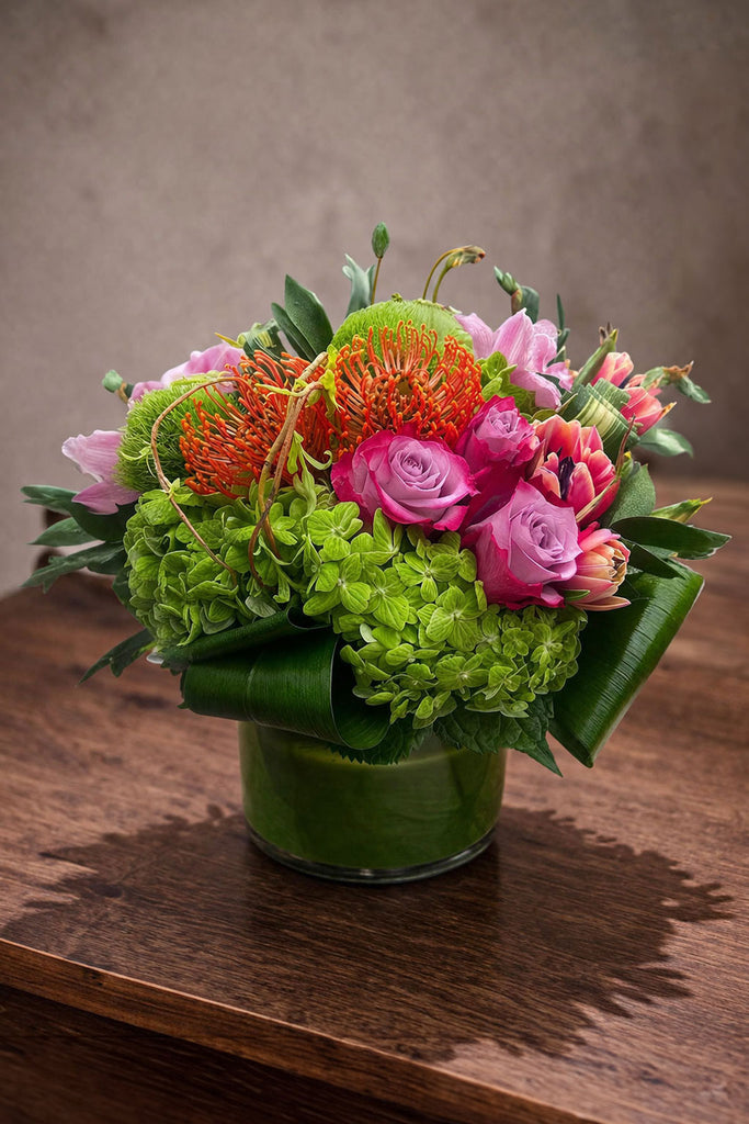 Beloved floral arrangement in a clear glass cylinder vase with pincushion protea, roses, green hydrangea, and colorful seasonal blooms.