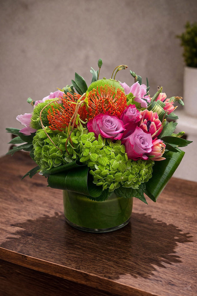 Beloved floral arrangement in a clear glass cylinder vase with pincushion protea, roses, green hydrangea, and colorful seasonal blooms.