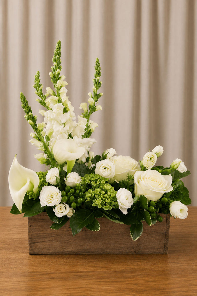 White and green floral arrangement with roses, calla lilies, snapdragons, lisianthus, green berries, and foliage in a rustic wooden box.