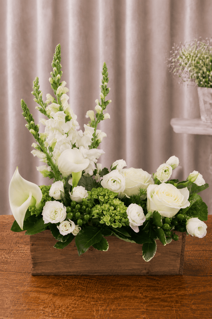 White and green floral arrangement with roses, calla lilies, snapdragons, lisianthus, green berries, and foliage in a rustic wooden box.