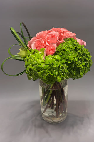 Modern vase arrangement with pink roses, green hydrangea, cymbidium orchid, bear grass loops, and tropical leaves in a clear glass cylinder vase.