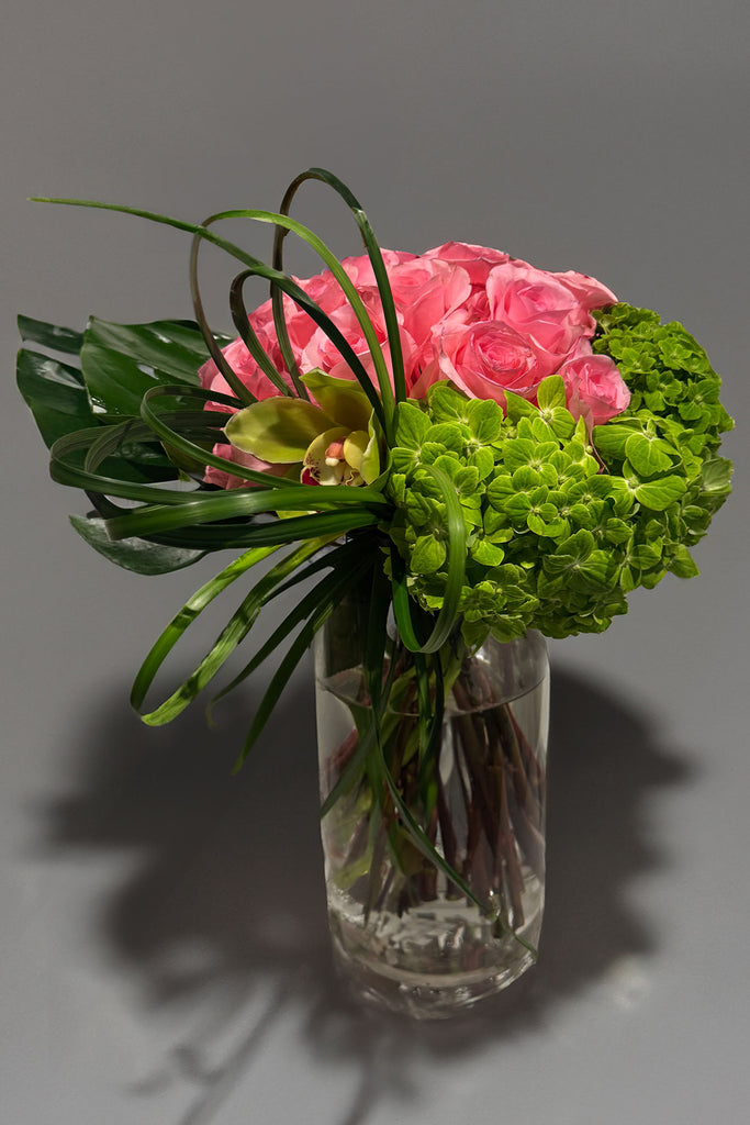 Modern vase arrangement with pink roses, green hydrangea, cymbidium orchid, bear grass loops, and tropical leaves in a clear glass cylinder vase.