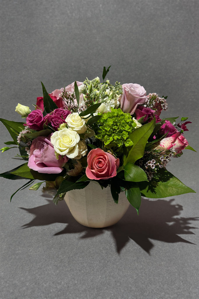 Vase arrangement with blush, lavender, and pink roses, accented with seasonal greenery and textures in a soft, rounded ceramic vase against a gray background.