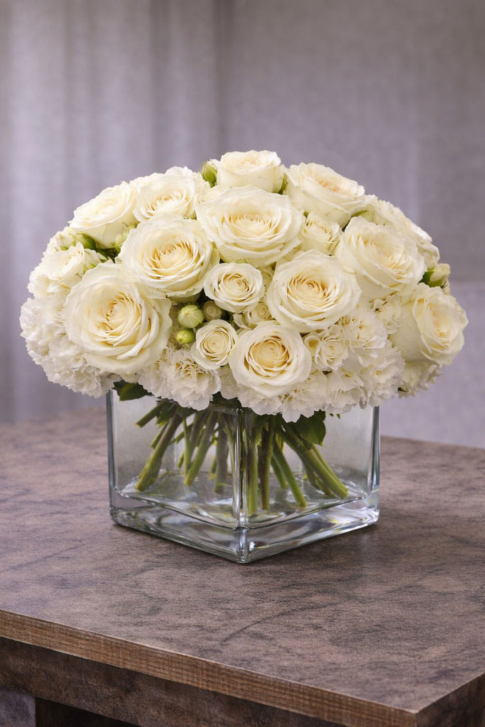 Blush floral arrangement in a clear glass cube vase with ivory roses, spray roses, and soft white blooms.