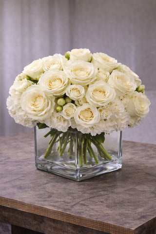 Blush floral arrangement in a clear glass cube vase with ivory roses, spray roses, and soft white blooms.