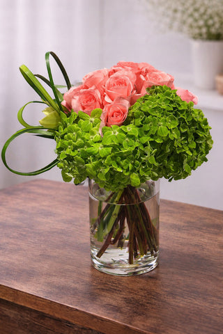 Pink roses and green hydrangea floral arrangement in a clear glass vase on a wooden table.