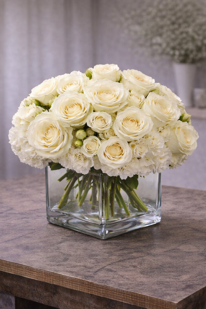 Blush floral arrangement in a clear glass cube vase with ivory roses, spray roses, and soft white blooms.