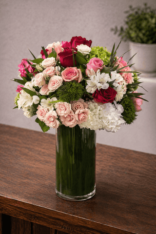 Blush Royale floral arrangement with blush, ivory, and red roses, white blooms, and hydrangea in a clear glass cylinder vase with green leaf lining.