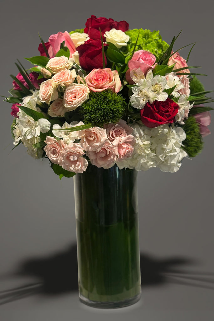 Tall glass vase arrangement with red and blush roses, hydrangea, cymbidium orchids, and layered greenery in a rounded, lush design.