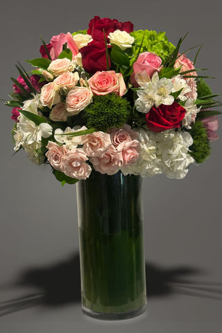 Tall glass vase arrangement with red and blush roses, hydrangea, cymbidium orchids, and layered greenery in a rounded, lush design.