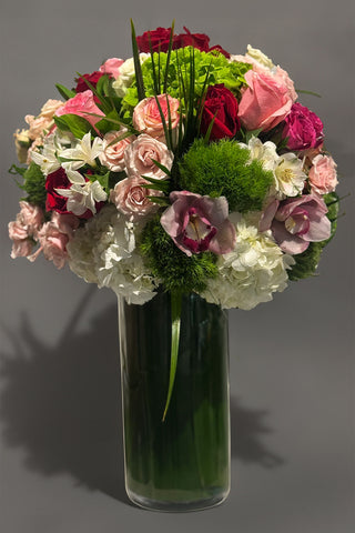 Tall glass vase arrangement with red and blush roses, hydrangea, cymbidium orchids, and layered greenery in a rounded, lush design.