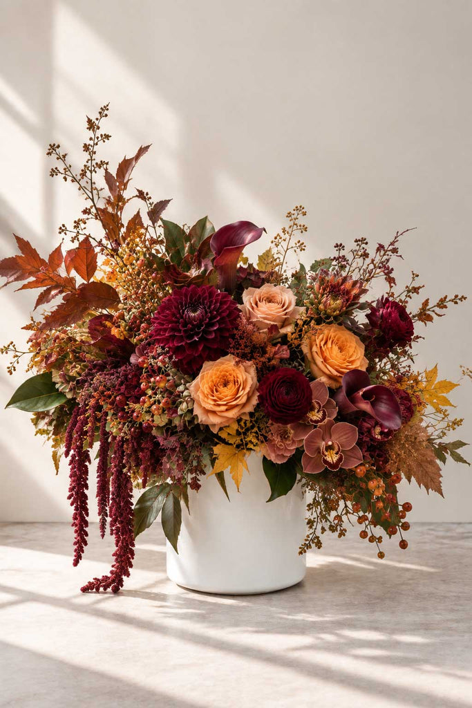Bordeaux Garden floral arrangement with burgundy dahlias, peach roses, calla lilies, cymbidium orchids, amaranthus, berries, and fall foliage in a white ceramic vase.