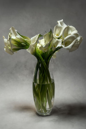 Clear glass vase with fresh white calla lilies arranged simply and elegantly, displayed against a soft gray background.