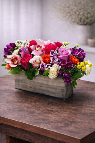 Cheerful floral arrangement in a rustic wooden box with roses, cymbidium orchids, colorful seasonal blooms, and fresh greenery.