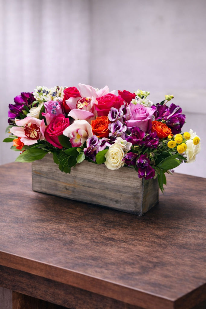 Cheerful floral arrangement in a rustic wooden box with roses, cymbidium orchids, colorful seasonal blooms, and fresh greenery.
