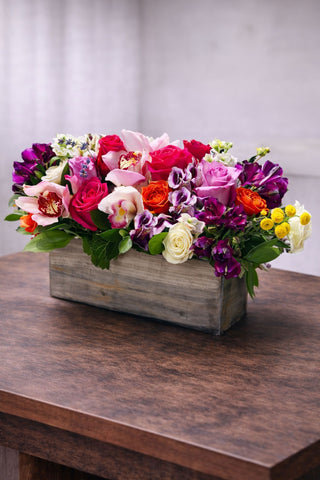 Cheerful floral arrangement in a rustic wooden box with roses, cymbidium orchids, colorful seasonal blooms, and fresh greenery.