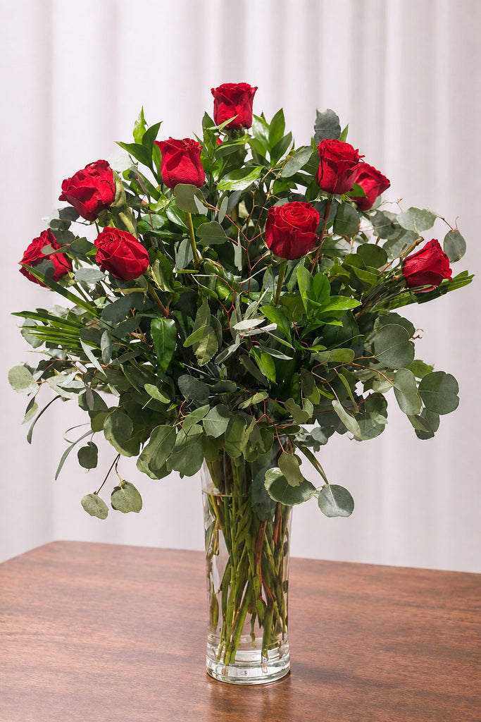 One dozen red roses with eucalyptus and lush greenery arranged in a clear glass vase on a wooden table.