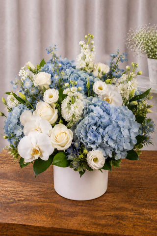 White and blue floral arrangement with blue hydrangeas, white roses, white orchids, and greenery in a white ceramic vase on a wooden table.