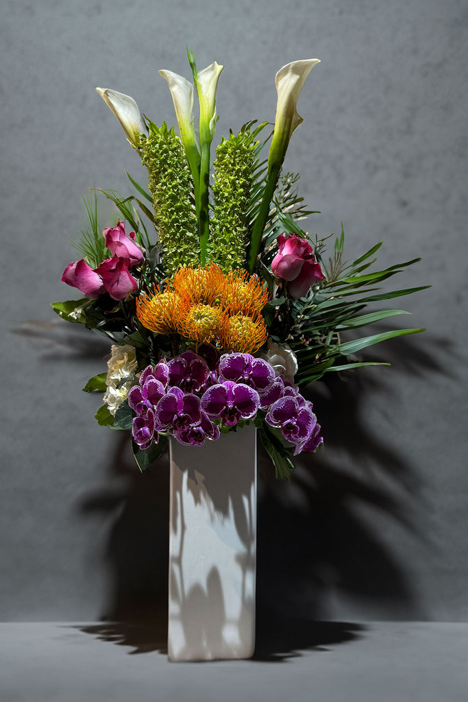 Tall modern vase arrangement with white calla lilies, orange pincushion proteas, purple orchids, pink roses, and tropical greenery.