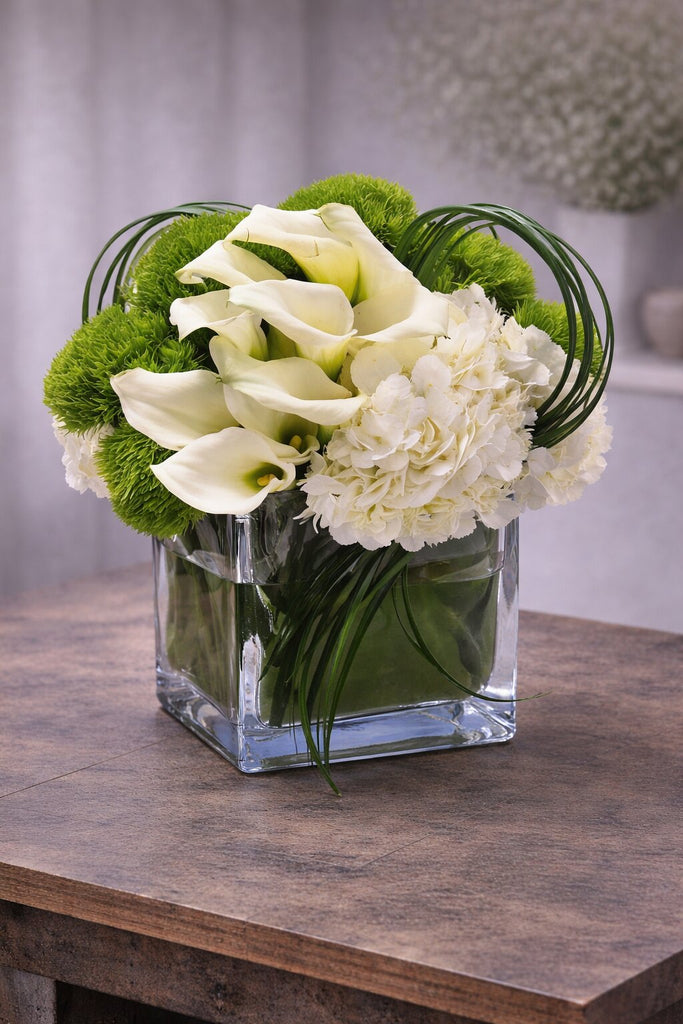 Dazzling floral arrangement in a clear glass cube vase with white calla lilies, white hydrangea, green dianthus, and looping bear grass.