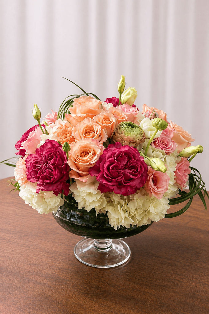 Soft pink and ivory floral arrangement with roses and hydrangea in a glass compote on a wooden table with natural light