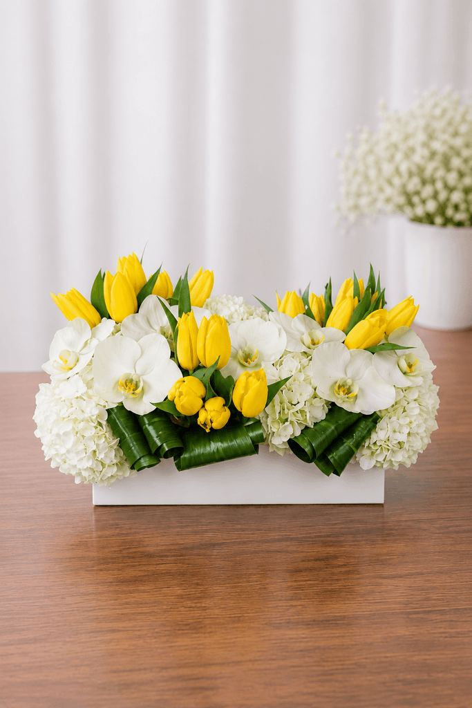 Yellow tulip and white orchid arrangement with white hydrangea and glossy green foliage in a white rectangular container.
