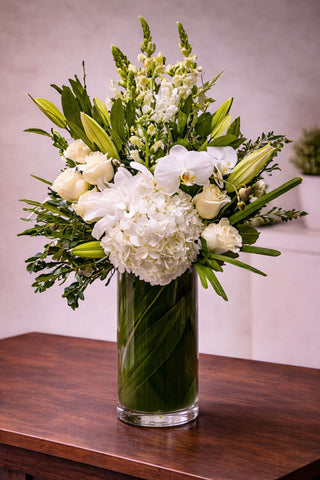 Everly floral arrangement in a tall glass vase wrapped with green ti leaves, featuring white hydrangea, roses, orchids, lilies, and seasonal blooms.