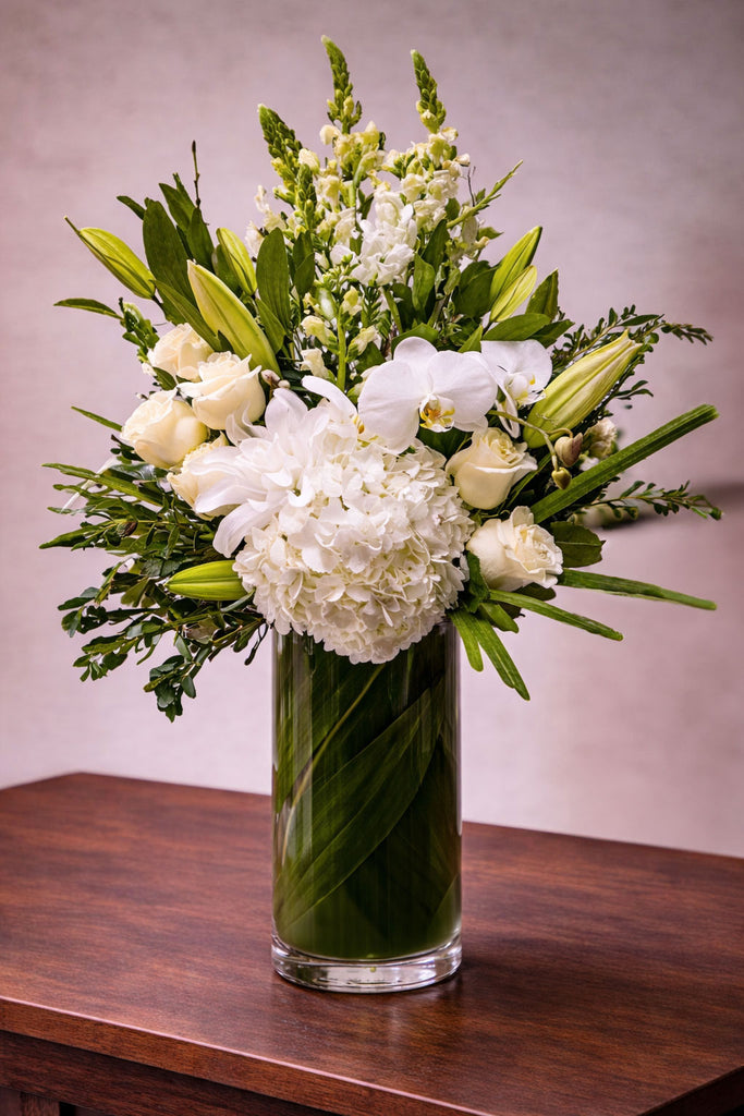 Everly floral arrangement in a tall glass vase wrapped with green ti leaves, featuring white hydrangea, roses, orchids, lilies, and seasonal blooms.