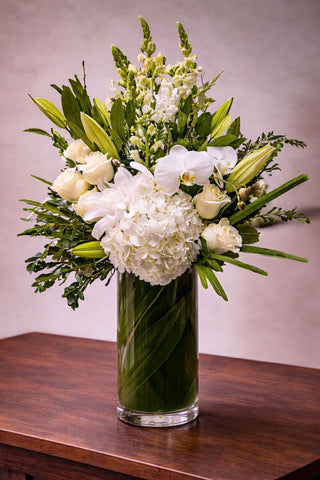 Everly floral arrangement in a tall glass vase wrapped with green ti leaves, featuring white hydrangea, roses, orchids, lilies, and seasonal blooms.