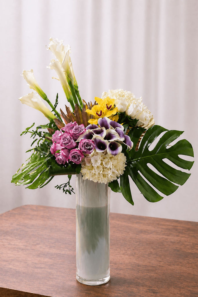 Tropical floral arrangement with white calla lilies, pink roses, yellow orchids, white hydrangea, purple mini calla lilies, and monstera leaves in a tall glass vase.