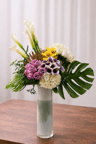 Tropical floral arrangement with white calla lilies, pink roses, yellow orchids, white hydrangea, purple mini calla lilies, and monstera leaves in a tall glass vase.