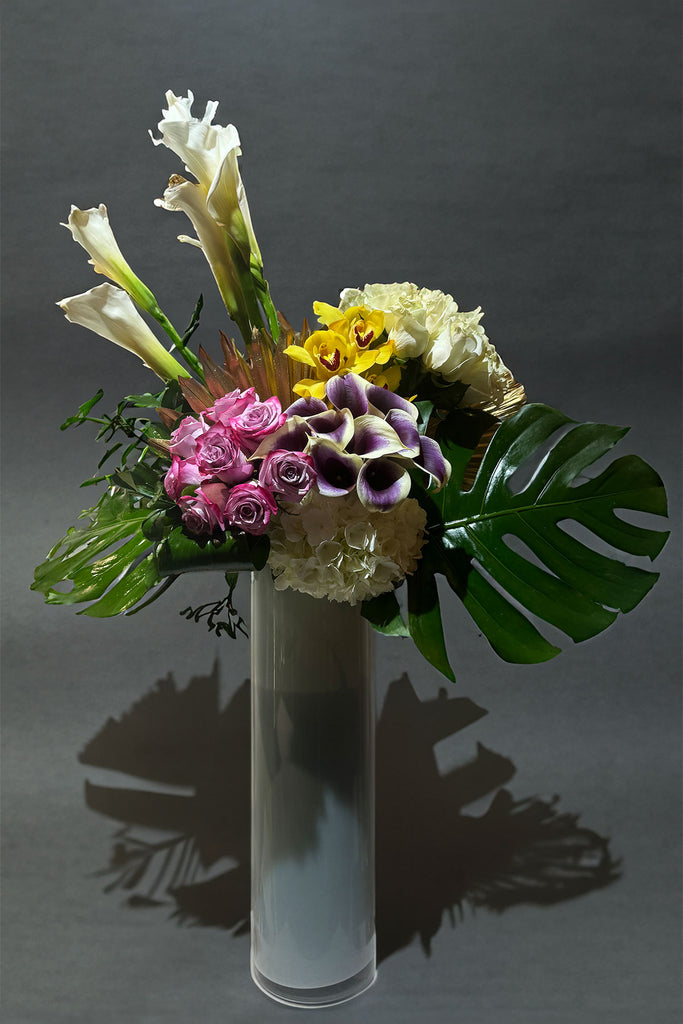 Tall vase arrangement with calla lilies, yellow orchids, roses, hydrangea, and monstera leaves, styled with artistic and tropical elegance.
