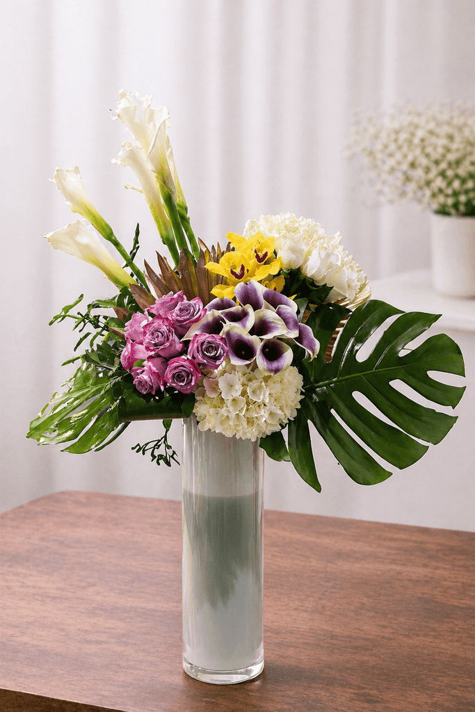 Tropical floral arrangement with white calla lilies, pink roses, yellow orchids, white hydrangea, purple mini calla lilies, and monstera leaves in a tall glass vase.