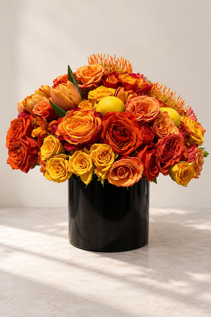 Golden Dawn floral arrangement with orange, amber, and yellow roses, pincushion blooms, and citrus accents in a black vase.