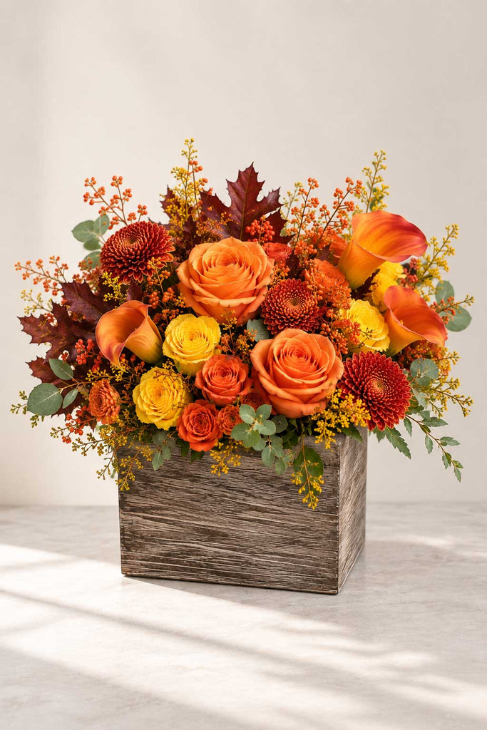 Golden Harvest floral arrangement with orange and yellow roses, burnt-orange calla lilies, rust mums, eucalyptus, berries, and fall foliage in a rustic wooden box.