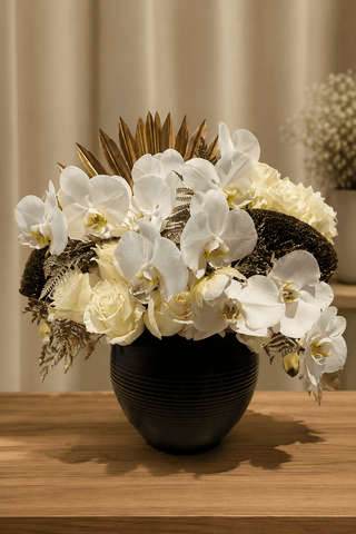 White orchid and ivory rose arrangement with golden botanical accents in a black vase on a wooden table.