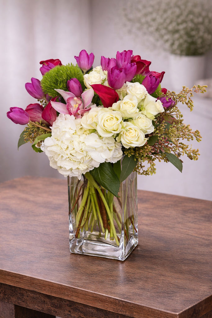 Hello Gorgeous floral arrangement in a clear glass vase with white hydrangea, ivory roses, pink tulips, cymbidium orchid, and seasonal accents.