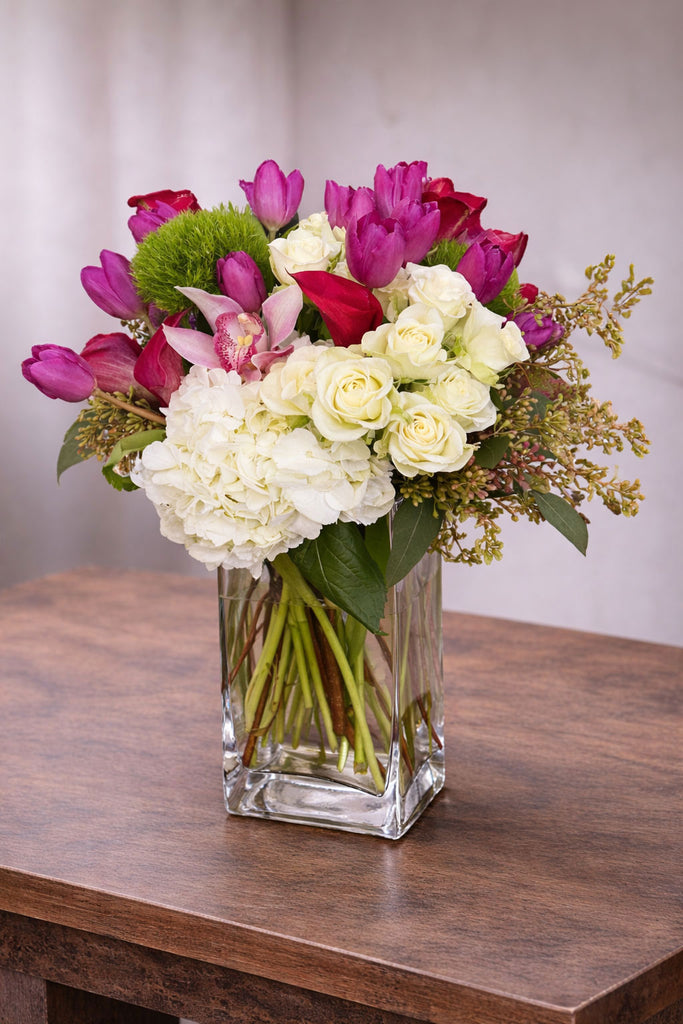 Hello Gorgeous floral arrangement in a clear glass vase with white hydrangea, ivory roses, pink tulips, cymbidium orchid, and seasonal accents.