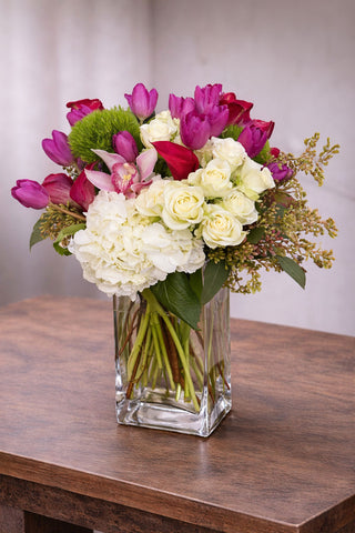 Hello Gorgeous floral arrangement in a clear glass vase with white hydrangea, ivory roses, pink tulips, cymbidium orchid, and seasonal accents.