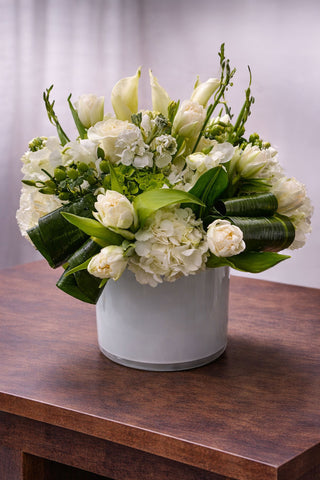 Minty Delight floral arrangement in a white glass vase with white calla lilies, hydrangea, tulips, roses, and fresh green accents.